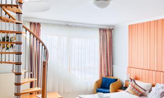 A bright room with a large bed and wooden floor. A spiral staircase leads to an upper floor, and the window lets in a lot of light.