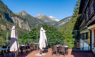A terrace with chairs and tables, surrounded by mountains and trees. The sky is clear and blue.