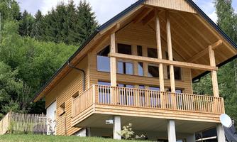 A modern wooden house with a balcony and large windows. It stands on a meadow, surrounded by trees.