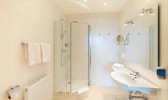 A modern bathroom with a shower and a sink. It is bright and minimally furnished, featuring white tiles and towels.