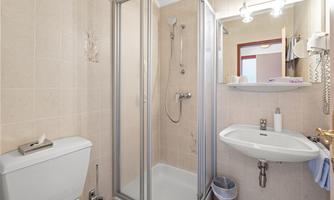 A modern bathroom with a shower, a sink, and a toilet. The walls are tiled in light colors, and there is a small shelf for hygiene items.