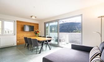 A modern living and dining room with a view of a lake and the mountains. The room is bright and welcoming furnished.