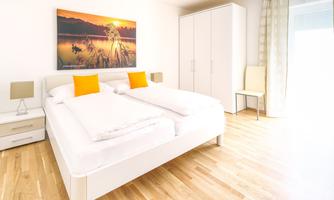 A modern furnished bedroom with a double bed and yellow pillows. A picture of a sunny landscape hangs on the wall, and the room has large windows for plenty of light.