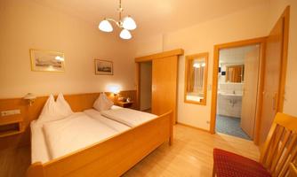 A cozy room with two single beds and bright wooden interiors. In the background, a bathroom is visible, accessible through a door.