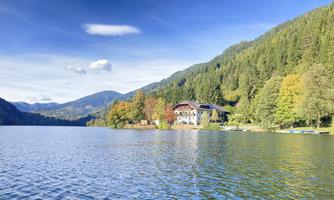 A picturesque lake surrounded by green mountains and colorful foliage. In the foreground, there is a beautiful building by the shore.
