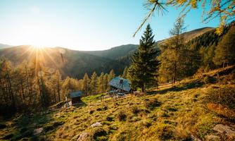 A picturesque mountain landscape with a cabin and trees. The sun rises behind the hills, bathing the scene in warm light.