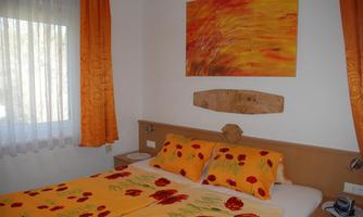 A cozy bedroom with a double bed and a colorful bedspread. The walls are decorated with orange curtains and a vibrant painting.