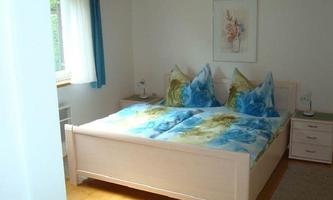 A cozy bedroom with a large bed and colorful pillows. The walls are light, and there are windows with curtains that let in daylight.