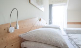 A modern bedroom with a wooden bed back and several pillows. There is a reading lamp and a large window area that lets in natural light.
