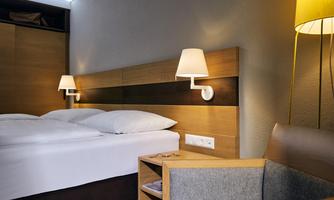 A modern hotel room with two beds and warm wooden tones. Next to the beds are wall lamps and a cozy armchair.