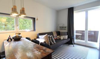 A modern living room with a cozy sofa and a wooden table.  
Large windows provide plenty of natural light and a view of the balcony.