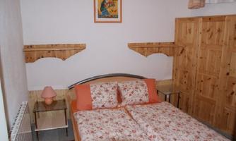 A cozy bedroom with a double bed and floral bedding. The walls are painted light, and there are wooden shelves as well as a small bedside lamp.