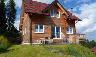 A beautiful wooden house with a red roof. It is situated amidst greenery and has large windows.