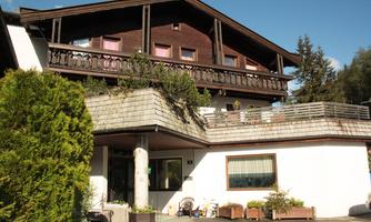 A charming wooden house with a balcony and green plants. The entrance is welcoming and the weather is sunny.