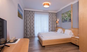 A modern bedroom with two beds, wooden furniture, and a large window. The walls are painted light and there are pictures on the walls.