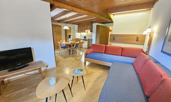 A bright and modern living room decor with a red and blue sofa. In the background, there is a dining table and a TV, surrounded by wooden ceilings and warm light.
