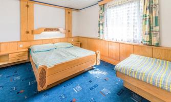 A simple bedroom with a double bed and a single bed. The walls are made of light wood, and there is a window with curtains.