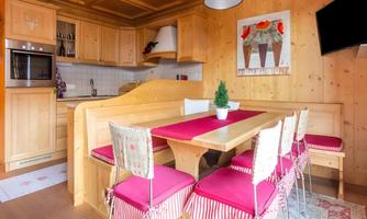 A cozy kitchen with wooden arrangements and a large dining table. The chairs are equipped with red cushions, and a plant is on the table.