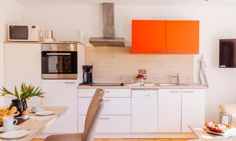 A modern kitchen with white cabinets and a striking orange upper cabinet door. It is well-equipped and has a cozy dining table in the foreground.