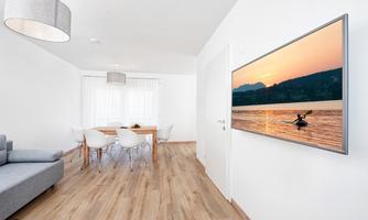 A modern living room with minimalist furnishings and a large TV displaying a landscape. The room has bright walls and wooden floors, with a dining area in the background.