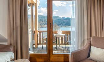 A bright room with a large glass door that leads to a balcony overlooking a lake and the surrounding mountains. The room is cozily furnished with a beige armchair and a table in the foreground.