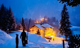 A winter scene with snow-covered trees and a illuminated house. Two people walk through the snow at twilight.