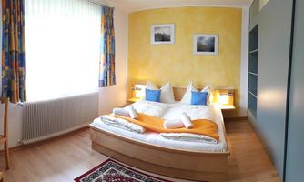 A cozy bedroom with a double bed and colorful cushions. The walls are painted in warm colors and large windows provide plenty of daylight.