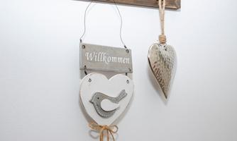 A heart-shaped welcome sign with a bird and a hanging heart-shaped decoration piece. Both elements are made of wood and have a rustic look.