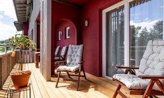 A cozy terrace with a wooden floor and two chairs. In the background, green plants and a red wall are visible.