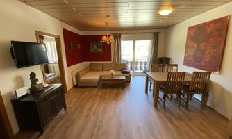 A bright living room with modern furniture and a wooden dining table. Large windows offer a view of the balcony.