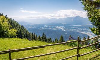 A beautiful view of the mountains with dense forests and a clear sky. In the foreground, there is a wooden fence that frames the picture.