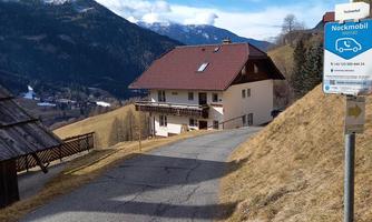 A picturesque mountain village with a two-story house on the hillside. In the background, the mountains and a clear sky can be seen.