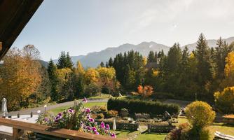 A picturesque mountain landscape with colorful autumn trees. In the foreground, plants bloom on a balcony, while the mountains in the background appear majestic.