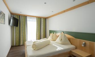 A modern hotel room with a double bed and two large pillows. The walls are light-colored and the curtains have a green pattern.