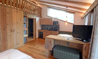 A cozy room with wooden details and a large window. The furnishings include a table, a television, and a small kitchenette.