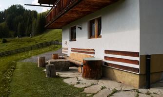 A rustic house in a green landscape with a wooden balcony. In front of the house, there are wooden furniture on a paved path.