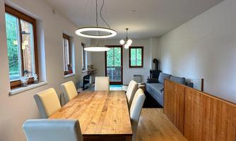 A modern dining area with a large wooden table and white chairs. The walls are light and there are large windows that let in a lot of light.