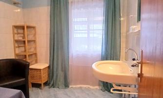A modern bathroom with light blue walls and a large window. There is a sink, a chair, and a wooden shelf.