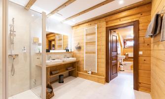 A modern bathroom with wooden paneling and a spacious shower. The room is bright and cozy, featuring a double sink and a window that provides natural light.