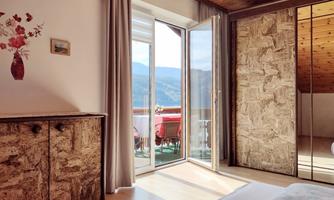 A cozy bedroom with a wood ceiling and a window that offers a view of the mountains. The decor is simple, featuring a dresser and a balcony in the background.