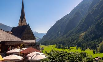 A picturesque landscape with tall mountains and a church in the foreground. The meadows are green and the sun is shining clearly in the sky.