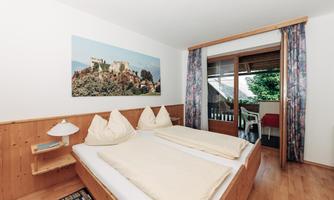A cozy bedroom with a double bed and wooden furniture. The windows offer a view of a balcony and plenty of daylight.