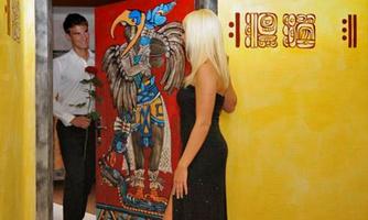 An elegant woman in a long black dress stands in front of a colorful door with an Aztec motif. A man with a rose looks from the other side of the door.