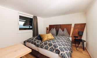 A modern bedroom with a large bed and elegant bedding. Next to the bed is a small table and a chair, and there is a window with a view outside.