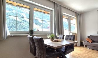 A modern dining area with a table and chairs, surrounded by large windows. Outside, a beautiful mountain landscape can be seen.