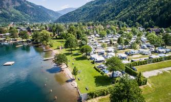 A picturesque campsite by the shore of a clear lake, surrounded by green mountains. Many motorhomes are scattered around the area, and some people are enjoying the water.