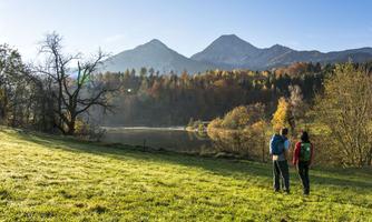 Two hikers are standing at the edge of a calm lake, looking at the surrounding mountains. The trees in the background display autumn colors under a clear blue sky.