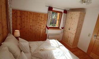 A cozy bedroom with wood paneling and a large window. The bedding is fluffed up and a wooden wardrobe stands in the room.