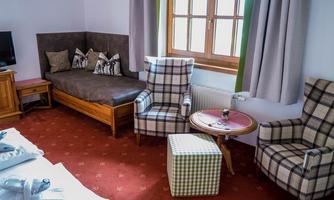 A cozy room with light curtains and two plaid armchairs. There is a large window and a wooden bench in the corner.