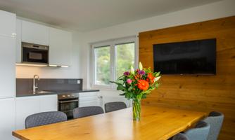 A modern kitchen with a large wooden table and colorful flowers in the center. A television hangs on the wall, and large windows provide plenty of light.